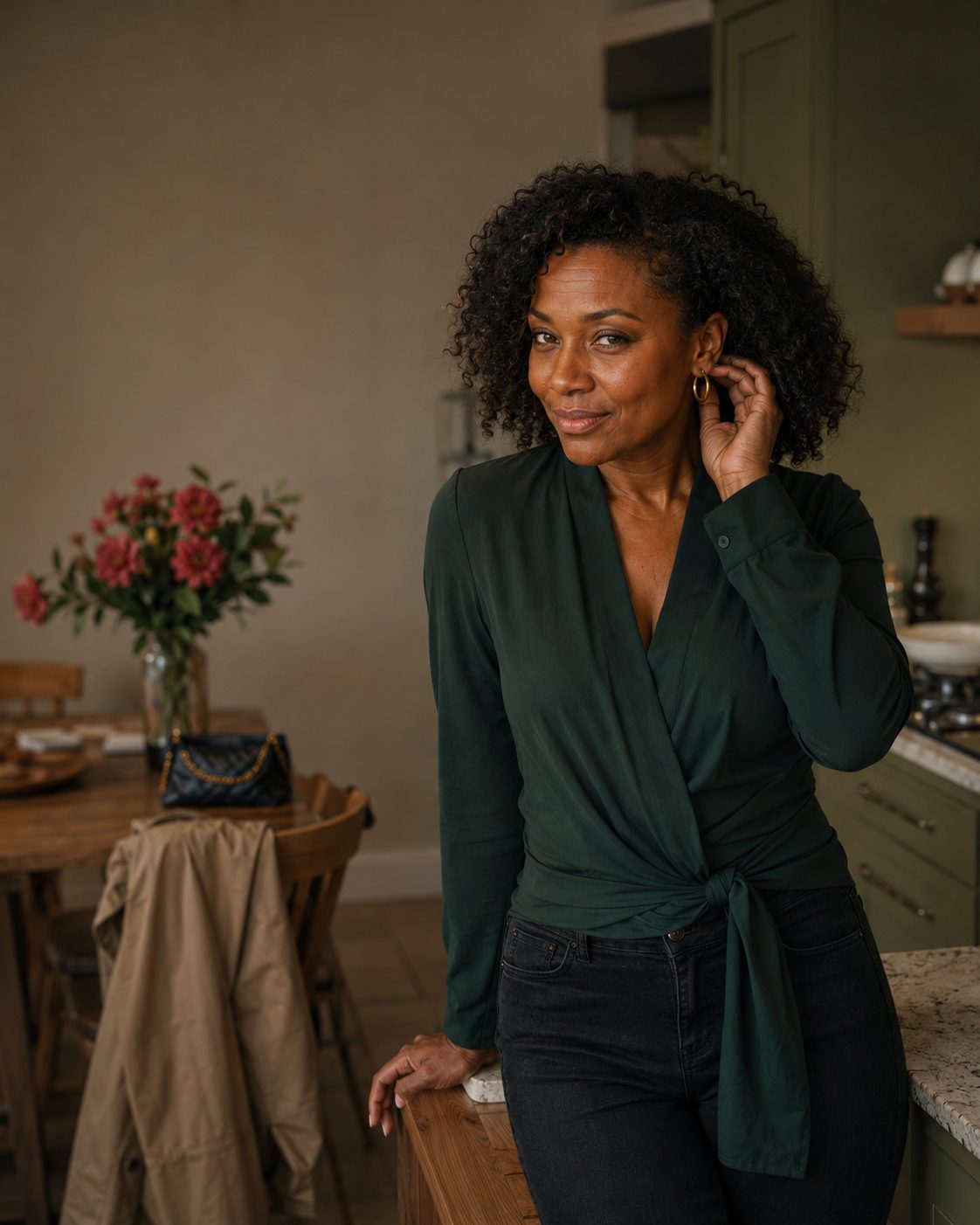 Woman with curly hair in a green wrap top standing in a kitchen beside a vase of flowers, tucking her hair back and smiling.