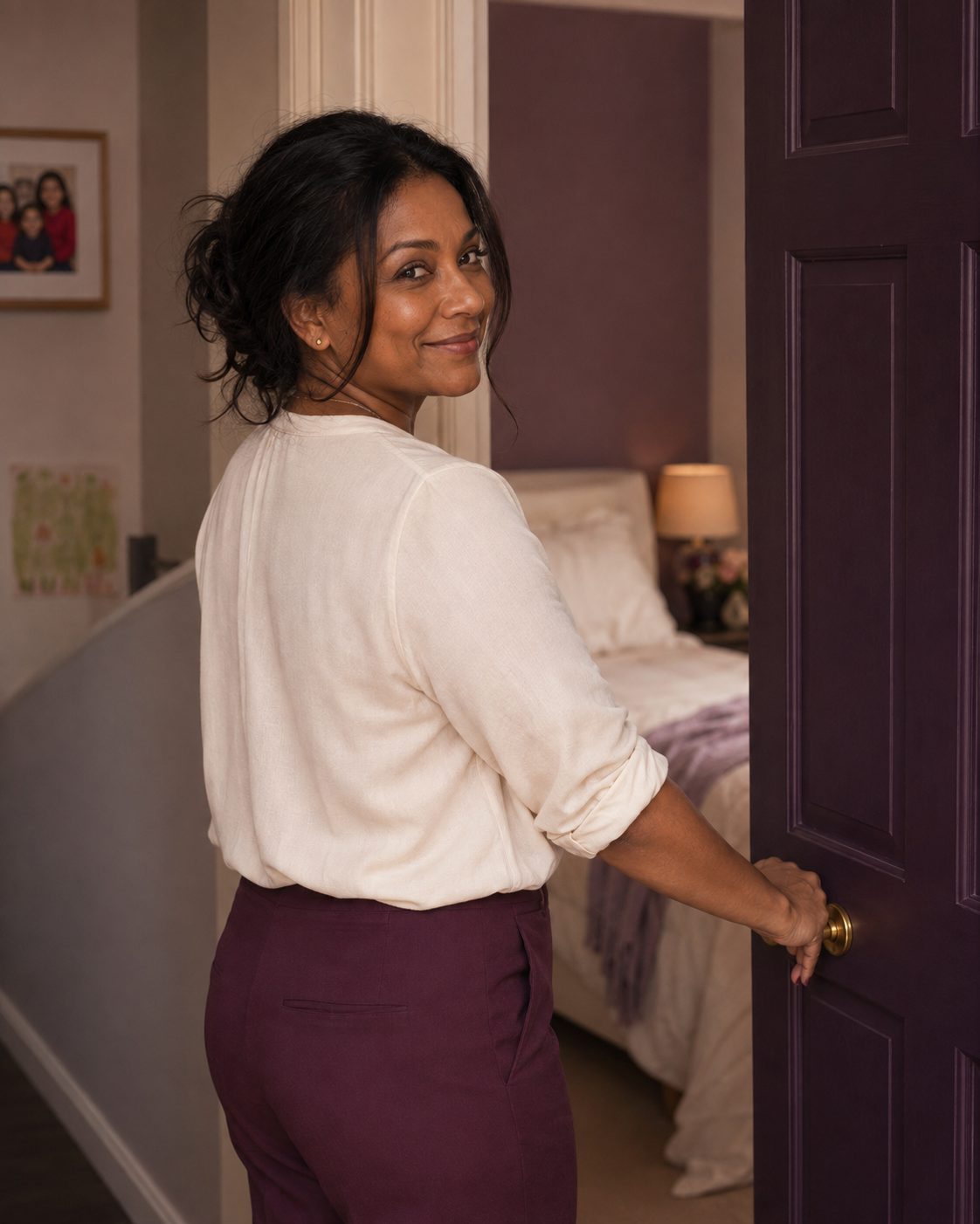 Woman in a cream blouse and plum trousers glancing back over her shoulder with a warm smile, opening a plum-painted bedroom door.