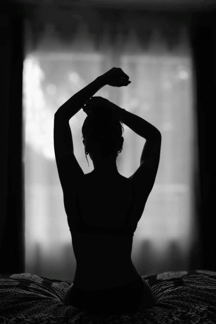 Black-and-white silhouette of a woman seen from behind, sitting on a bed with her arms raised in front of a bright window.
